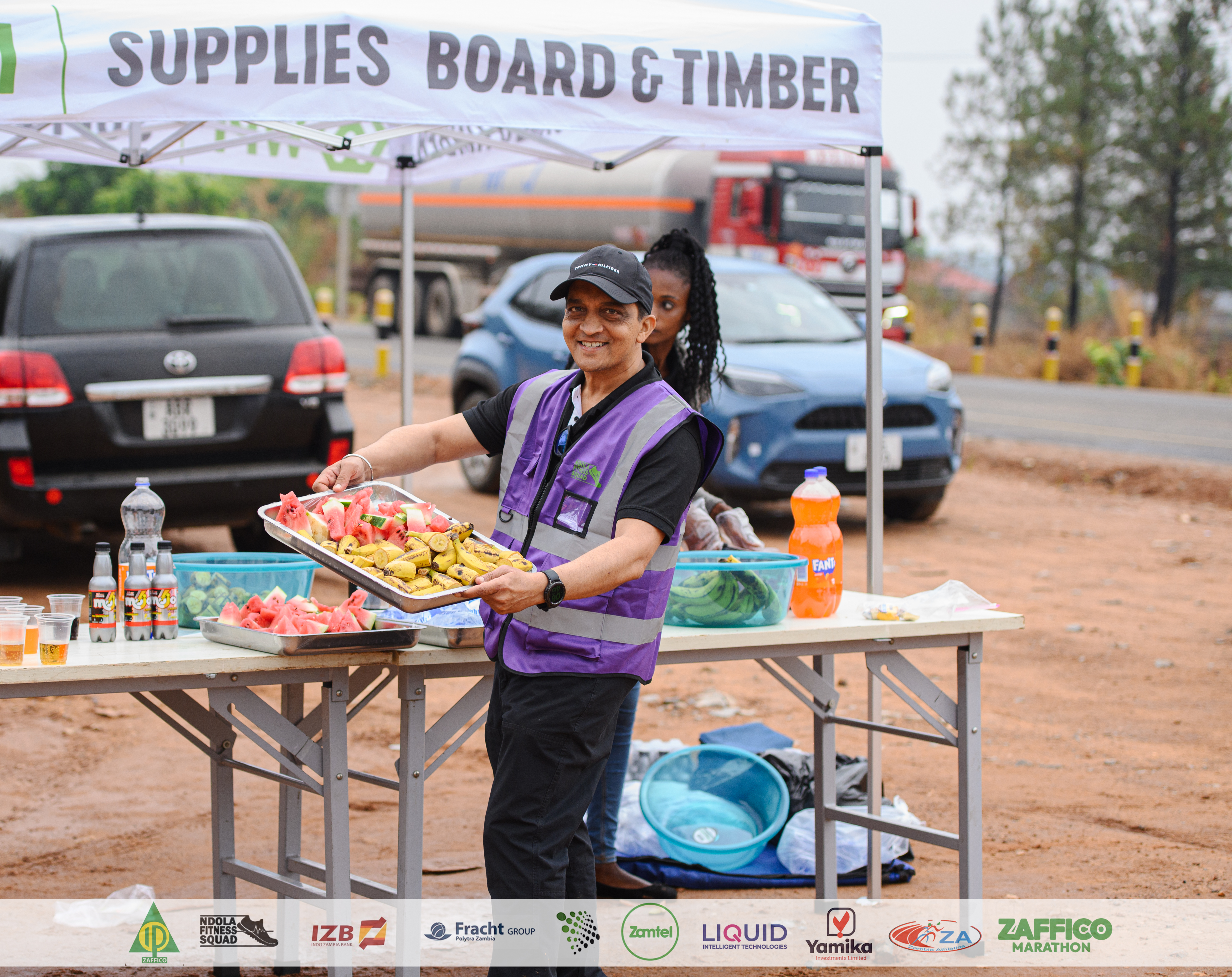 Volunteer in a purple vest offering a tray of fresh watermelon and bananas at a ZAFFICO Marathon aid station
