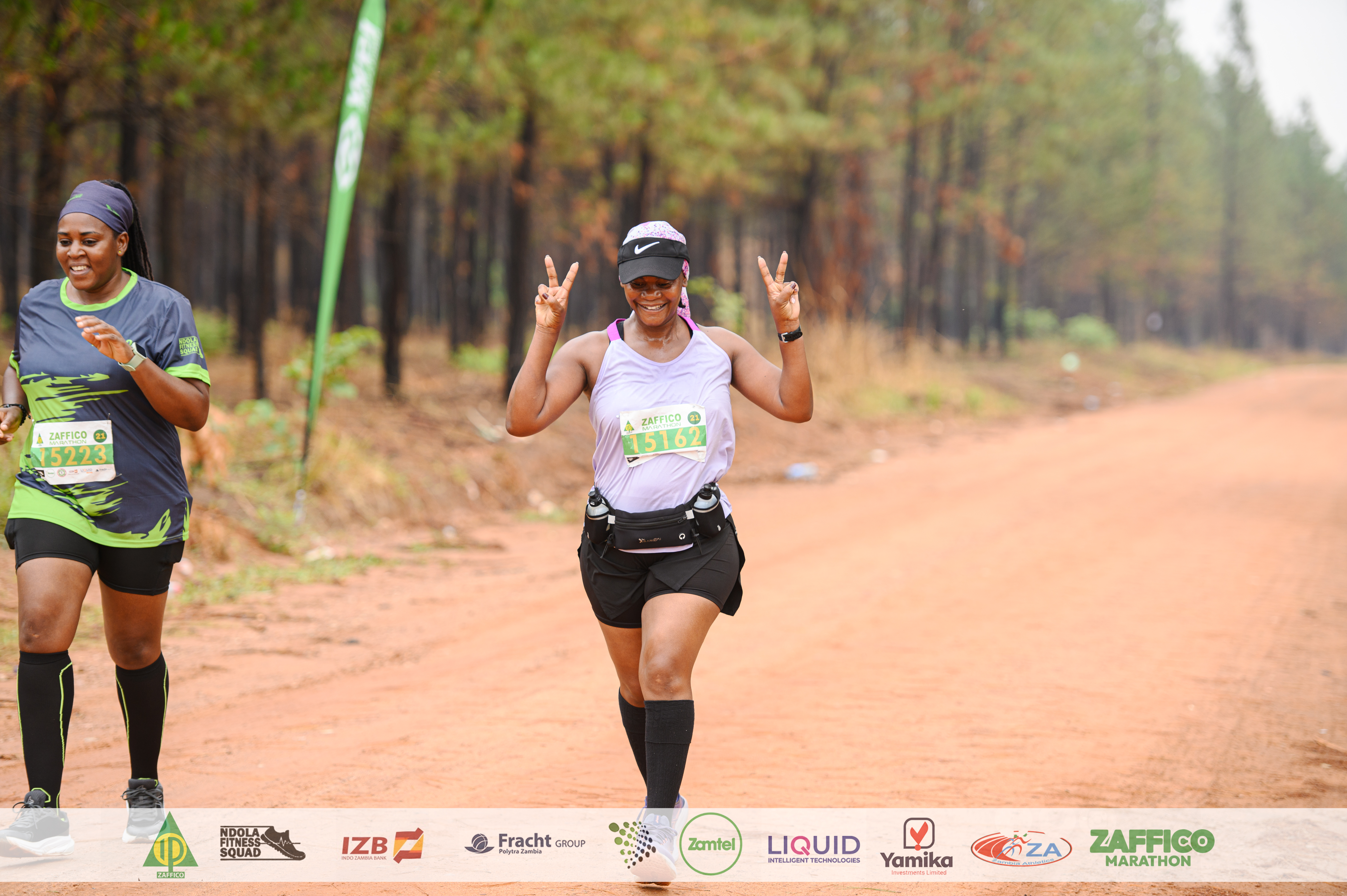 Runner in a lilac tank flashing double peace signs on a forest dirt road during the ZAFFICO Marathon